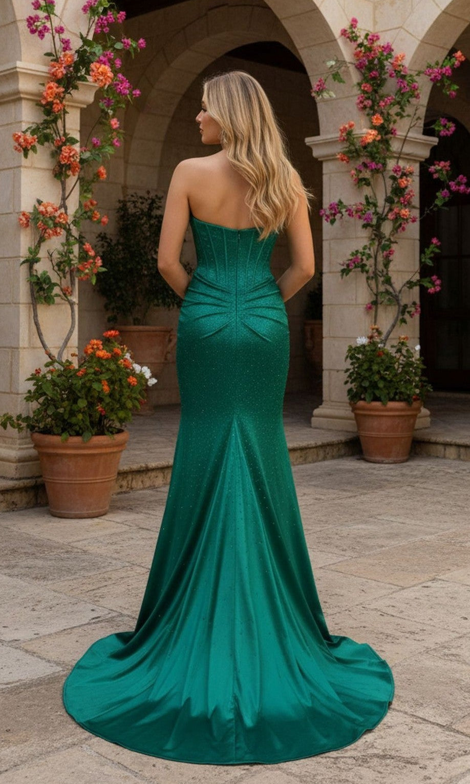 A woman with long blonde hair wears the PromGirl Exclusive 250216 fitted emerald green satin corset prom dress, standing outdoors on a stone patio and facing away from the camera.